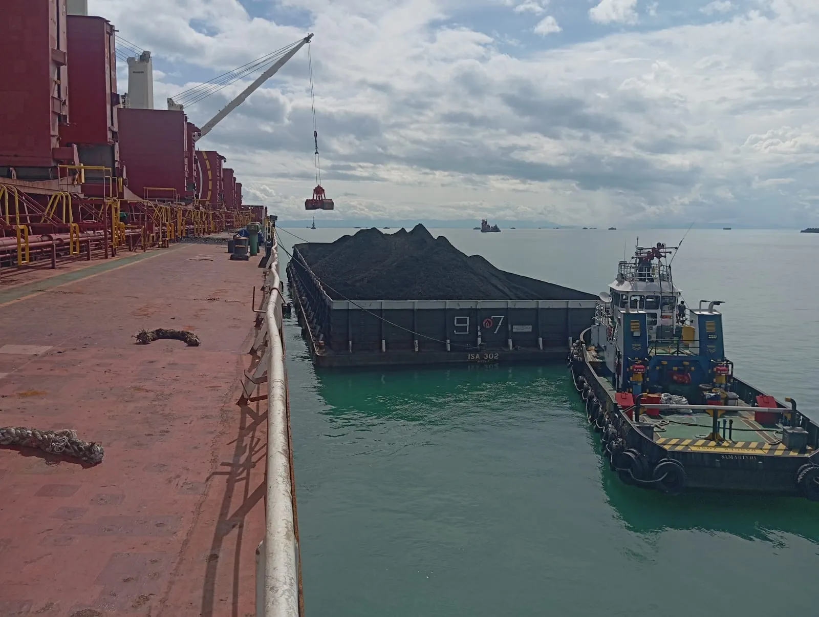 Tongkang bermuatan batu bara dan tugboat bersandar di sisi kapal besar di perairan lepas pantai